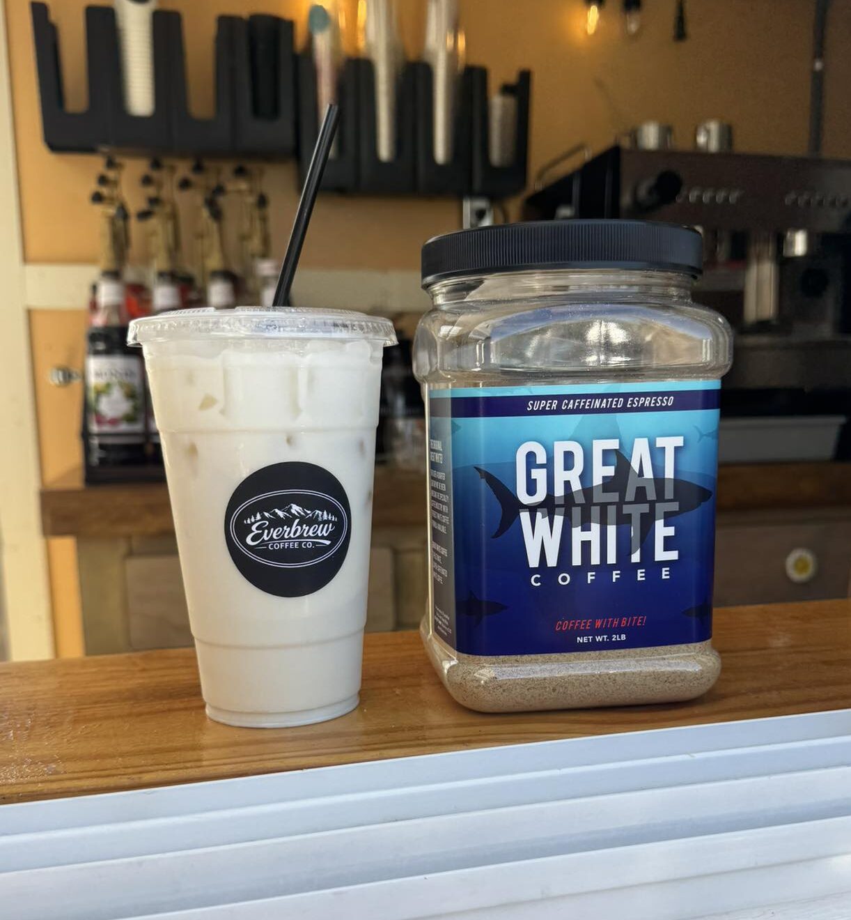 Iced white coffee in a clear plastic cup with lid and straw sitting on counter next to a clear container of "Great White" coffee.
