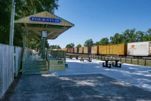 A Train passes by a covered stage area with a sign "Folkston Funnel" that sits adjacent to a paved seating area with concreate picnic tables