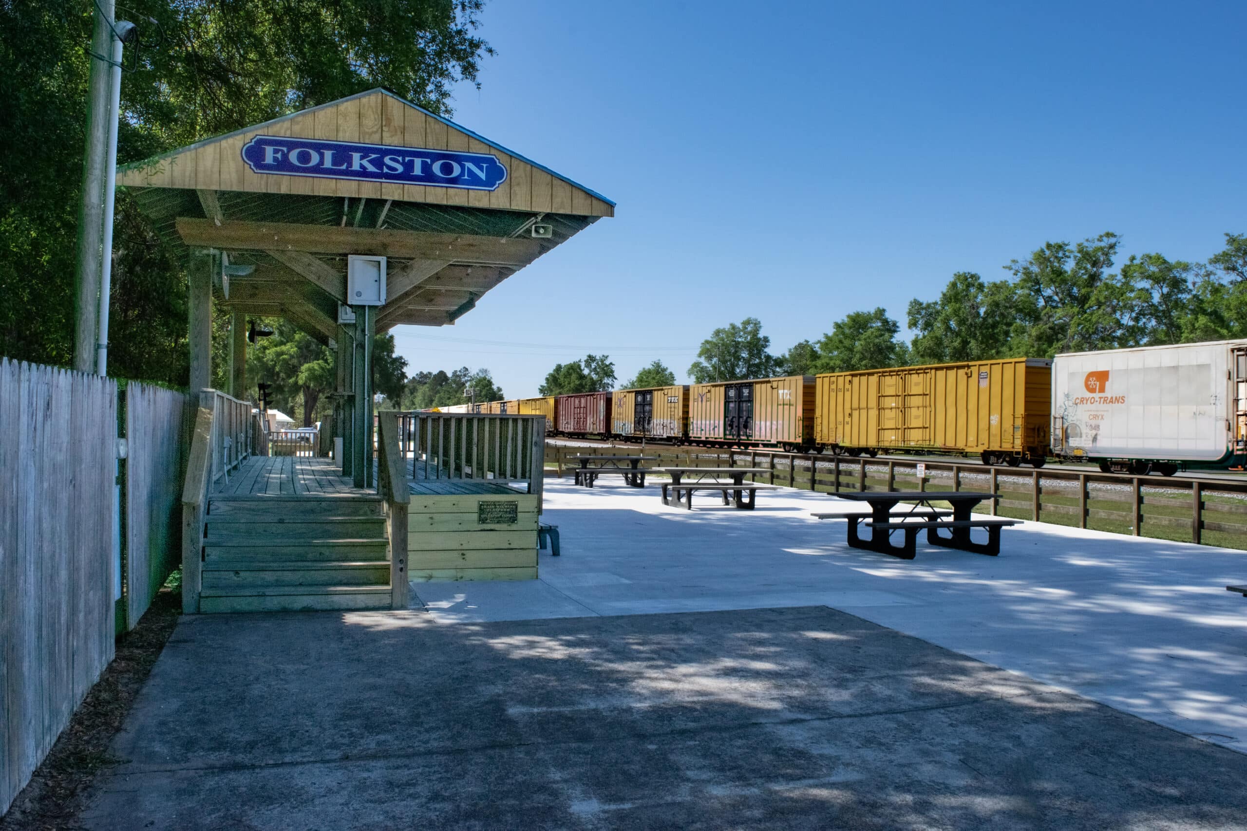 A Train passes by a covered stage area with a sign "Folkston Funnel" that sits adjacent to a paved seating area with concreate picnic tables