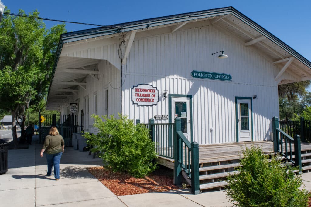 A woman walks along the sidewalk next to an old train depot building that says "Folkston, GA" Another sign tells us that this building is also the Okefenokee Chamber of Commerce that houses a Train Museum
