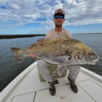 Captain Andrew Allen of Risen Tide Charter Co. sitting on a YETI cooler on the bow of a fishing boat and holding a large, fresh caught Grouper