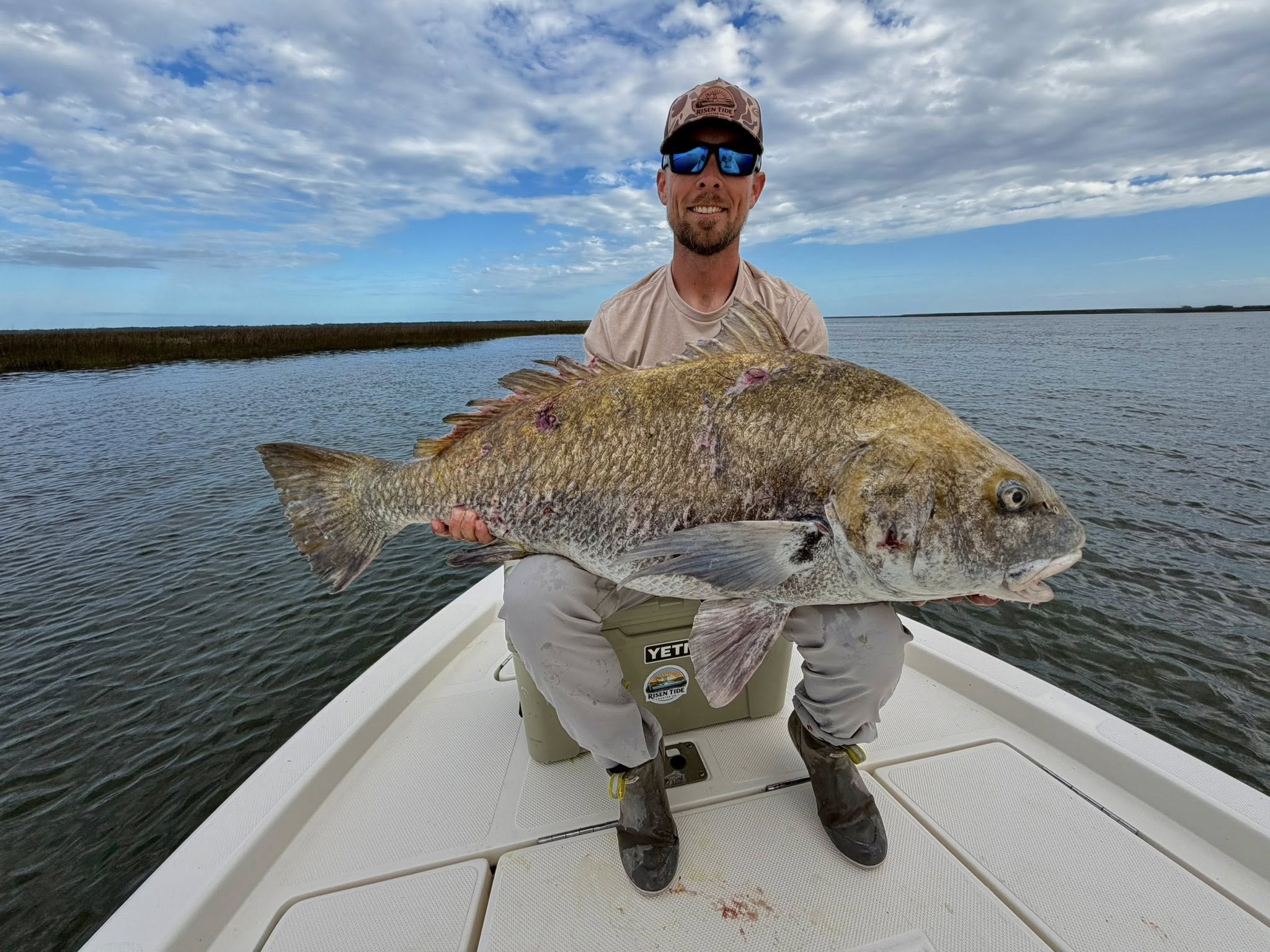 Captain Andrew Allen of Risen Tide Charter Co. sitting on a YETI cooler on the bow of a fishing boat and holding a large, fresh caught Grouper