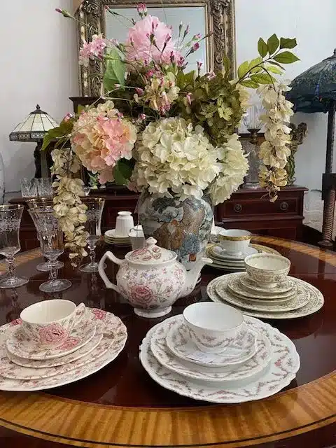 Vase of flowers on a round wooden table with various sets of fine china and crytal glasses with antique lamps and an ornate mirror on the wall behind