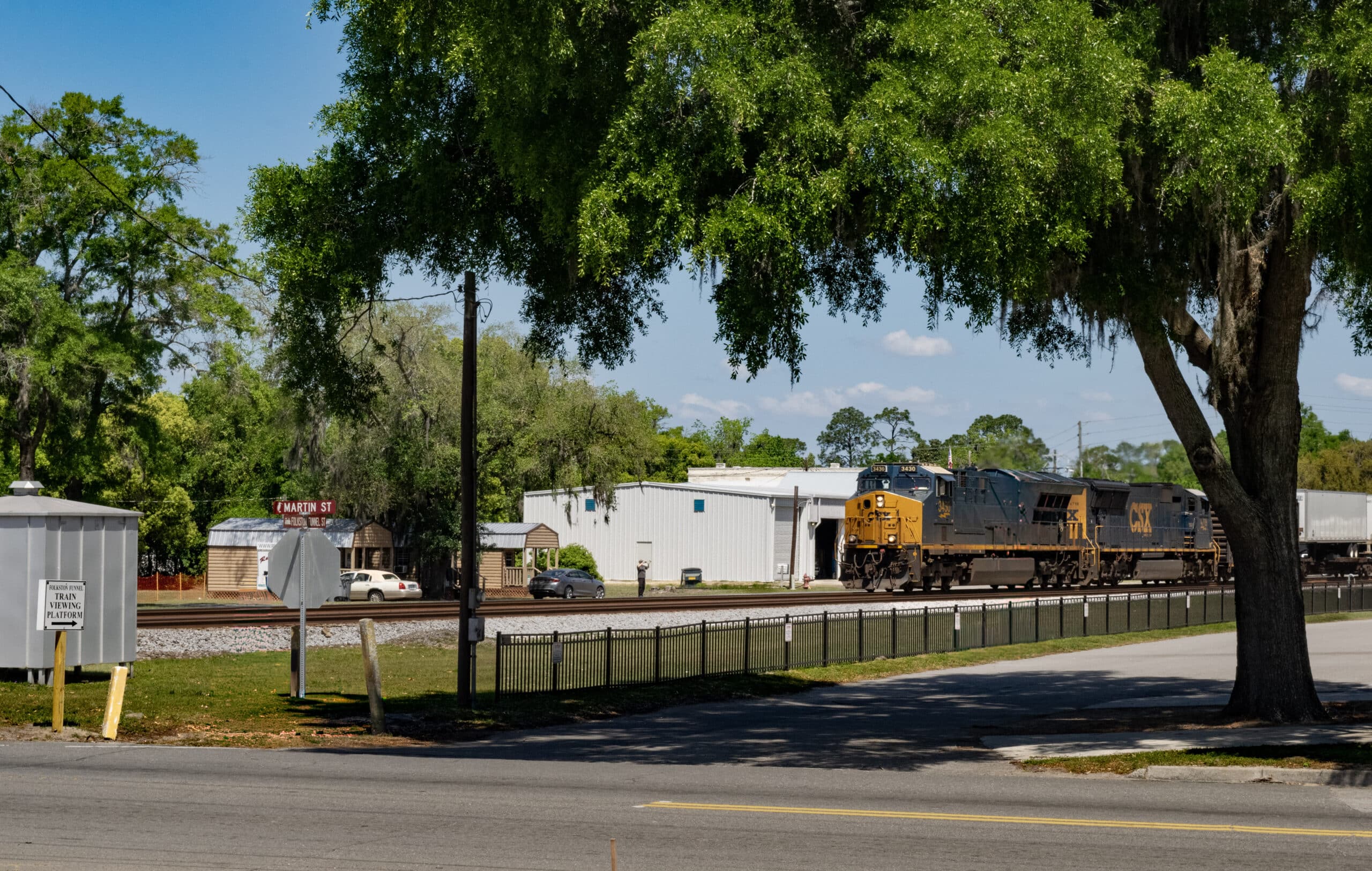A Tree provides a shady area over the sign for "Train Viewing Platform" as a train passes by the Folkston Funnel in Folkston, GA