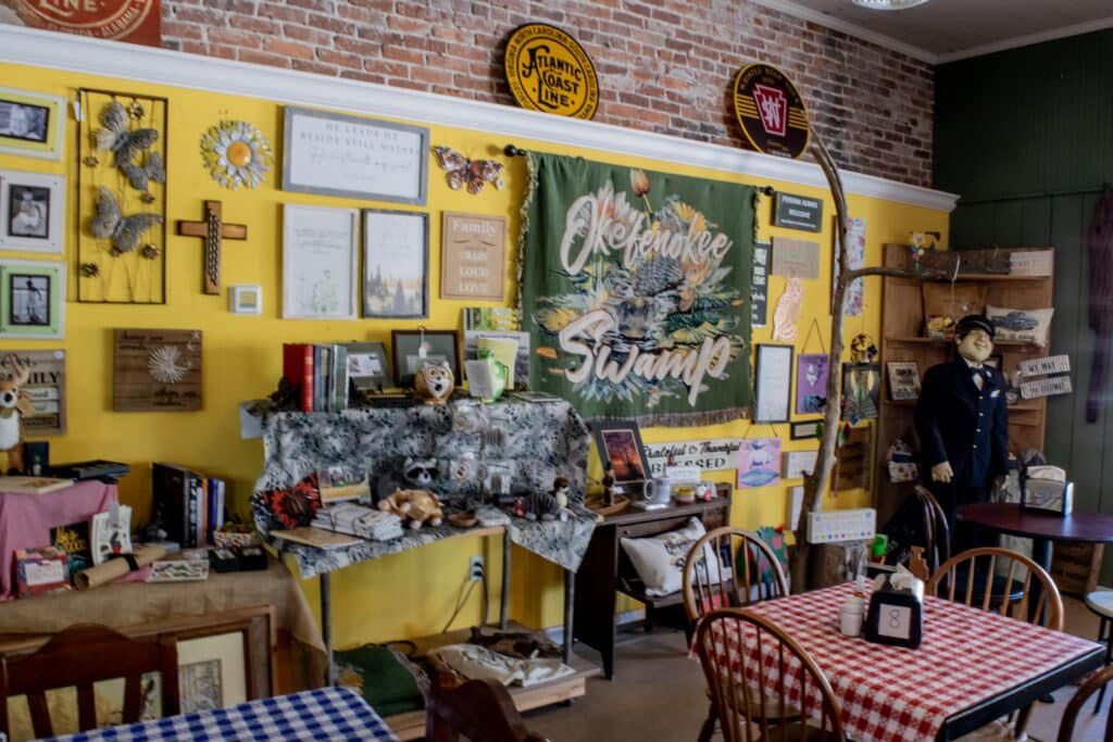 A blanket depicting the Okefenokee Swamp hangs along a wall covered with other rustic merchandise behind two dining tables with checkerboard tablecloths in the Whistlin Dixie Cafe in Folkston, GA