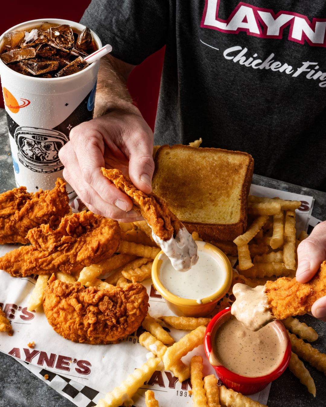 Close up of someone eating a Layne's Chicken Finger meal with crinkle-cut fries, Texas toast, an iced cola in Layne's branded Styrofoam cup, and two dipping sauces