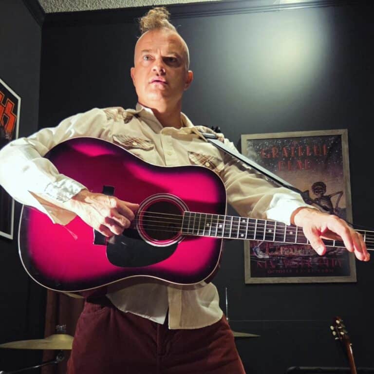 White man in a long-sleeved, collared white shirt, holding a black and red acoustic guitar and wearing his blonde hair in a mohawk.