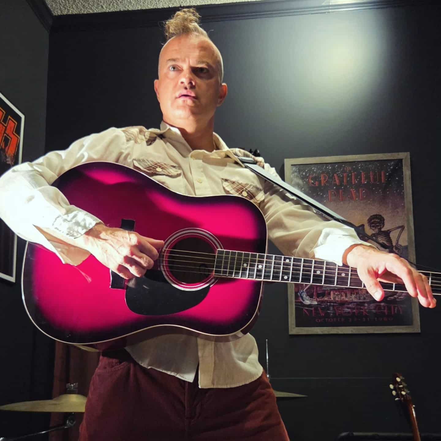 White man in a long-sleeved, collared white shirt, holding a black and red acoustic guitar and wearing his blonde hair in a mohawk.