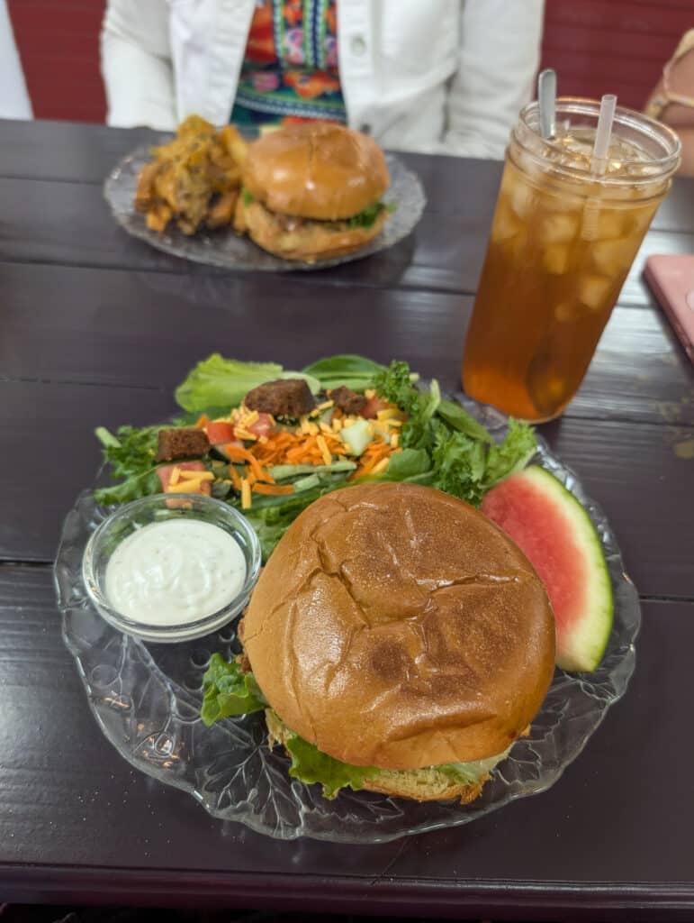 Sandwich on a brioche roll with a salad and dressing on a plate. A glass of iced tea on the side