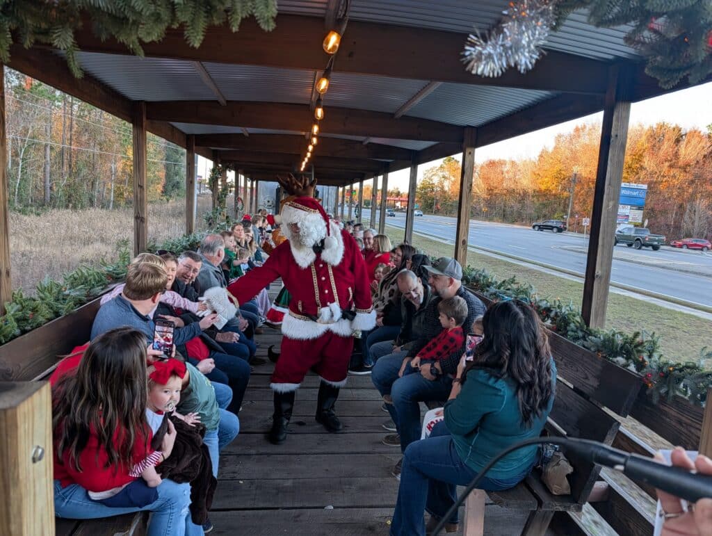 Santa walking down the aisle of an open-air train car speaking with families seated on wooden benches