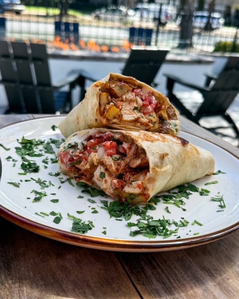 Burrito sliced in half on a white plate with cilantro sprinkles on a wooden table with Adirondack chairs and a firepit in the background.