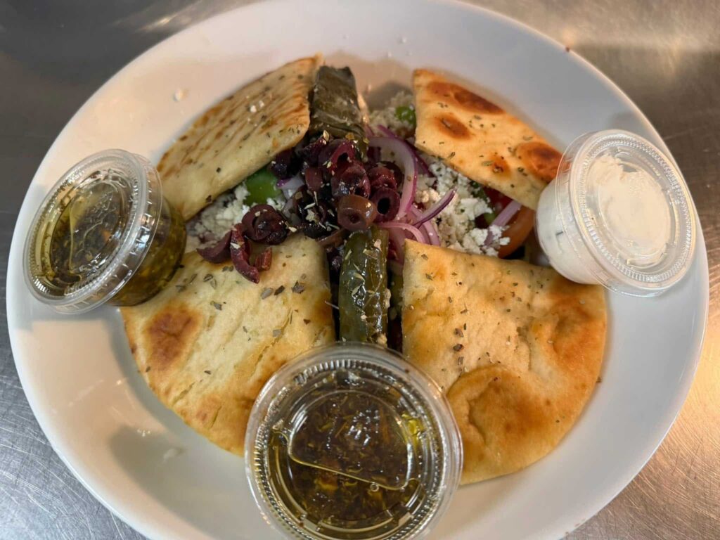Greek salad with four pita wedges and sides of Greek dressing and tzatziki sauce in plastic cups, all in a white bowl