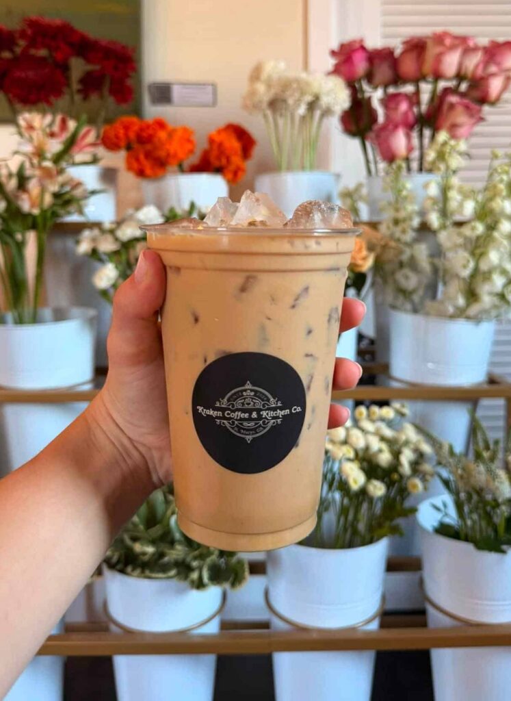 Hand holding a an iced coffee in a clear cup with Kraken Coffee branding with a display of flowers for sale in the background
