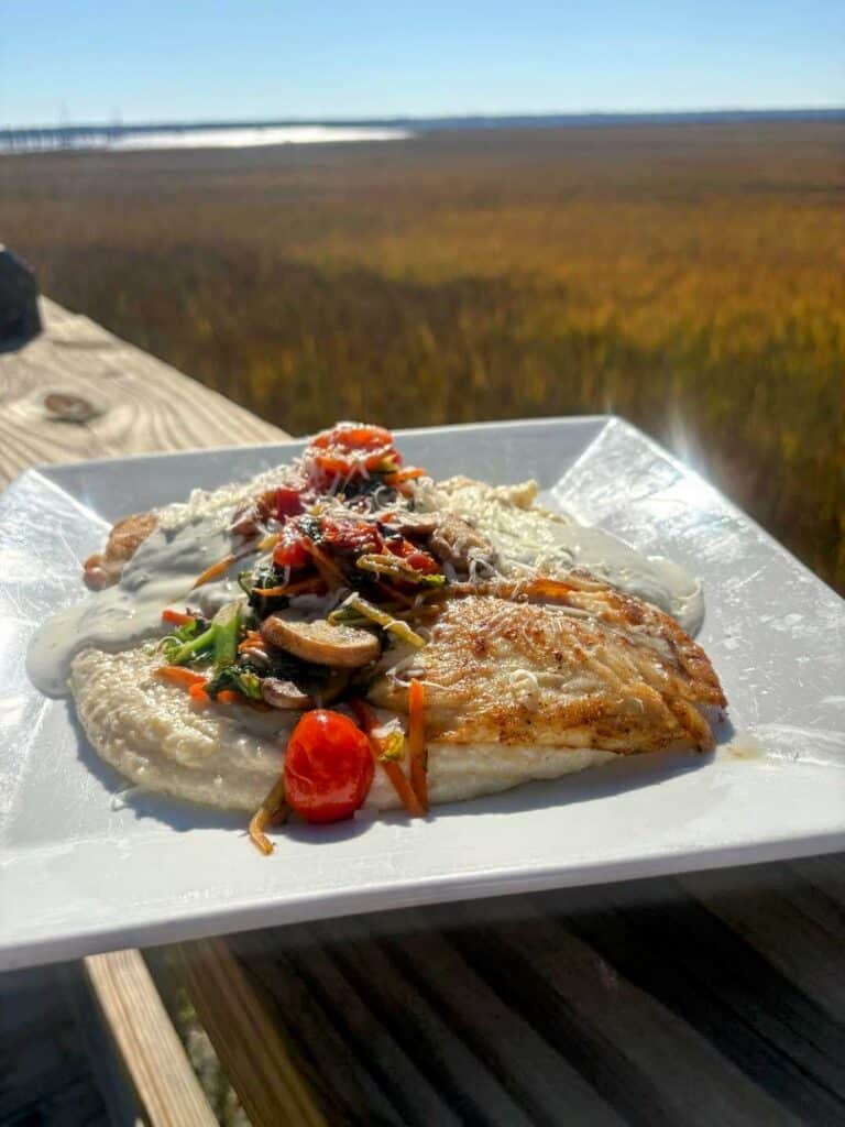 A plate of grilled Atlantic Tripletail served over grits with a mushroom cream sauce and cherry tomatoes sitting on a square white plate on a wooden deck overlooking marshland and river on a sunny day.