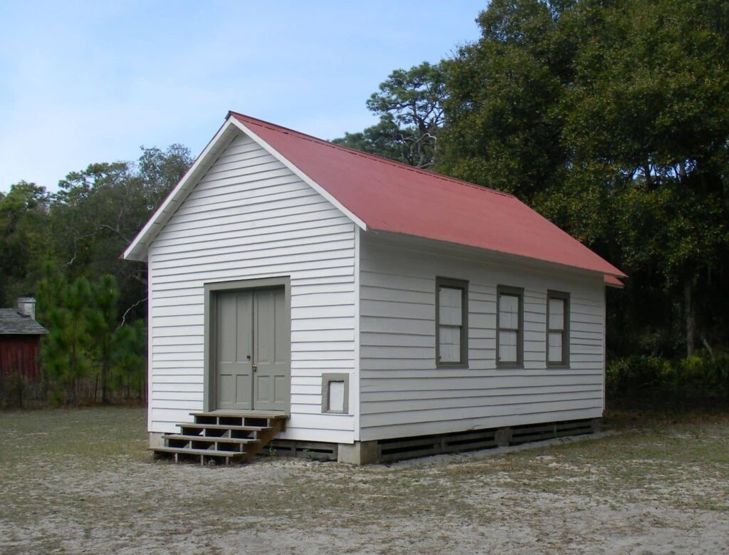 Small white shiplapped building with red a-frame roof and gray double doors on a sandy plot with trees in the background.