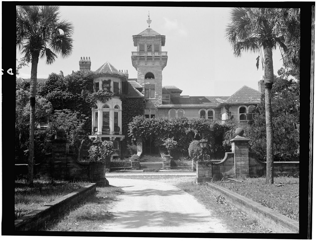 Black and white photo of palm tree-lined driveway approaching multi-story gilded age mansion taken in 1958.