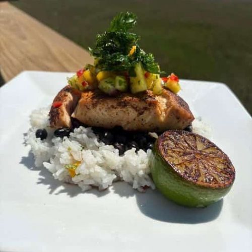 Blackend Mahi Mahi on a bed of black beans and white rice topped with pineapple mango salsa and parsley garnish and a half a lime that's been seared on the cut side--all on a white plate sitting on a wood railing with the salt marsh in the background